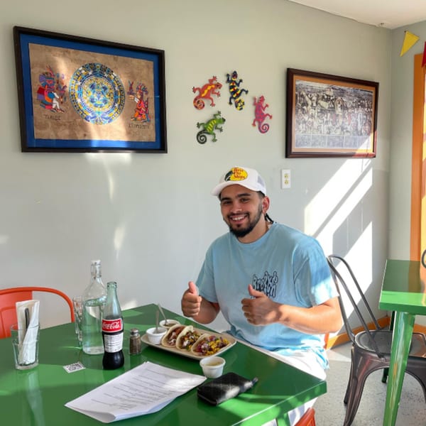 A man in a white cap and blue shirt gives thumbs up at a green table with tacos.