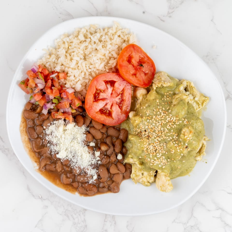 A white plate with chicken in green sauce, rice, beans, pico de gallo, and tomato slices.