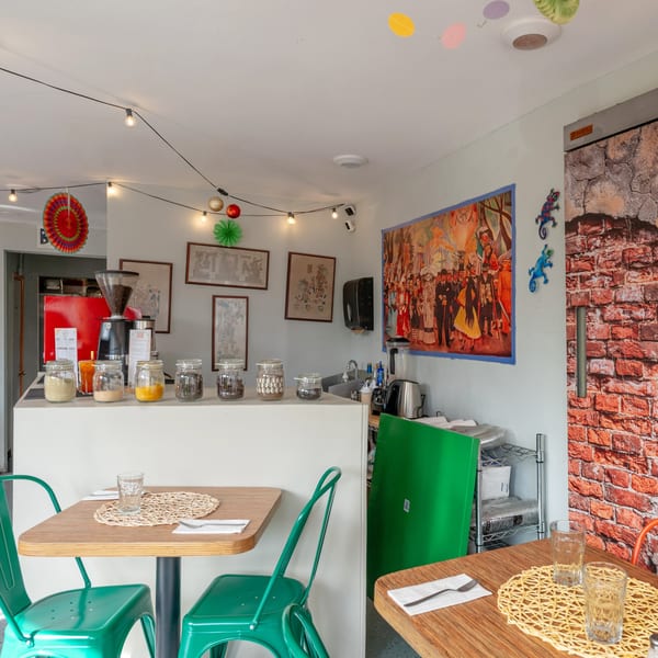 A vibrant restaurant interior with wooden tables, green chairs, a coffee bar, and colorful wall art.