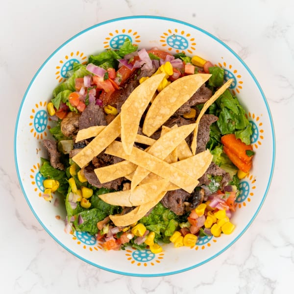 Overhead view of a colorful salad with steak, corn, tomatoes, and tortilla strips in a decorative bowl.