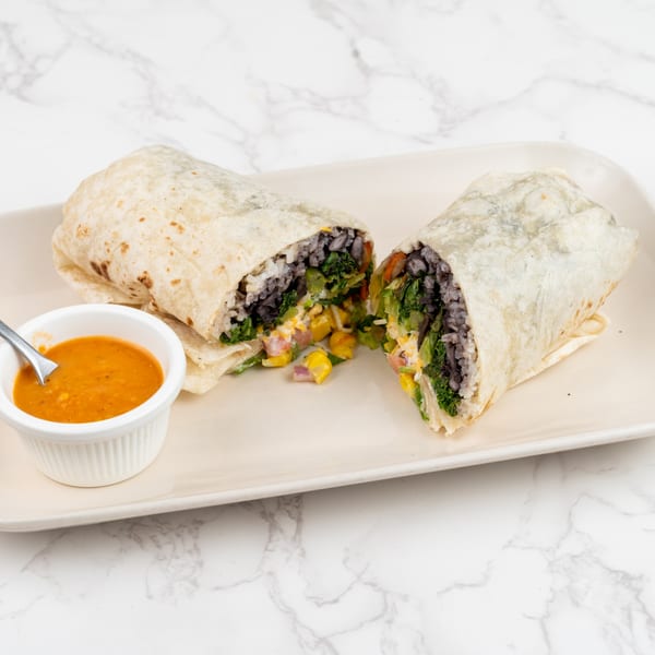 A large burrito, cut in half to show black beans, rice, corn, and greens, with a side of orange sauce.