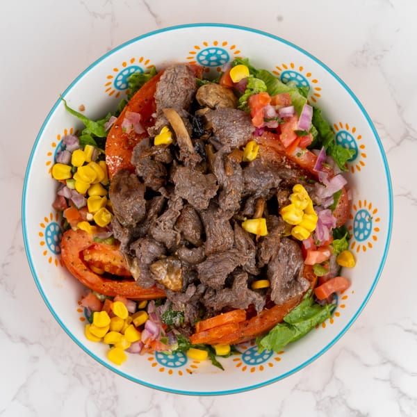 Overhead view of a colorful bowl of beef salad with corn, tomatoes, and lettuce.