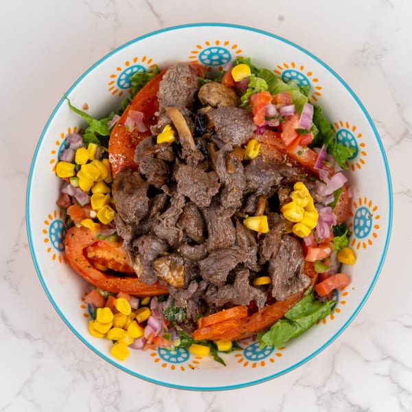 Overhead view of a colorful salad with sliced beef, corn, tomatoes, and red onions in a decorative bowl.